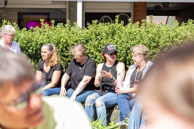 Gruppe af kvinder sidder udendørs og samtaler nær en hæk. Nogle har jeans på, andre solbriller. Solskinsdag.