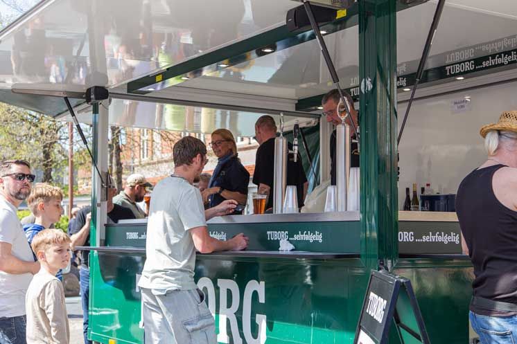 Folk bestiller øl på en udendørs Carlsberg-bar; grøn facade, solskinsdag.
