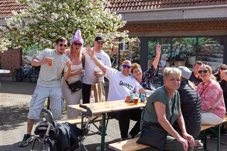 Gruppe af mennesker smiler, vinker og drikker ved et udendørs bord; solskinsdag.