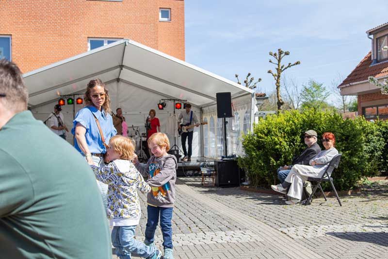 Udendørskoncert: Band på scenen, børn der leger, folk kigger på, solskinsdag.