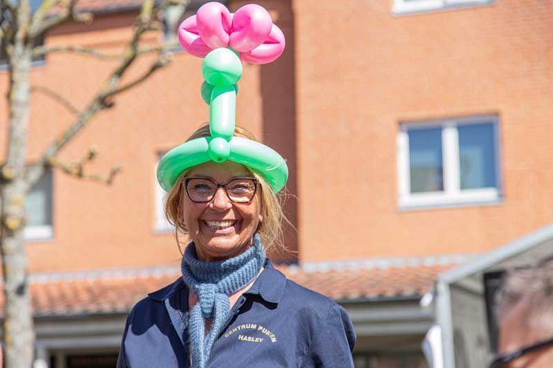 Kvinde iført en grøn og lyserød ballonhat smiler udendørs foran en bygning.