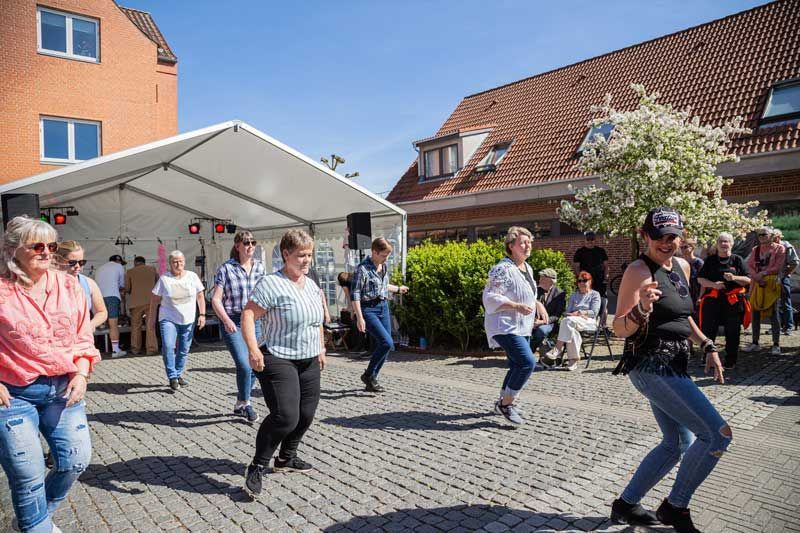 Folk danser udendørs foran en scene under et telt på en solskinsdag. Brostensbelagt jord og murstensbygninger i baggrunden.