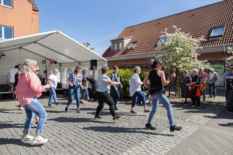 Folk danser på en bytorv; hvid teltscene i baggrunden, solskinsdag.