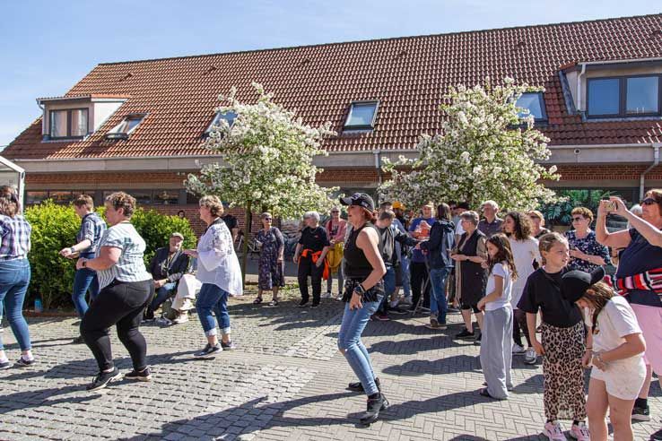 Folk danser udendørs på en solskinsdag foran en bygning med rødt tegltag.