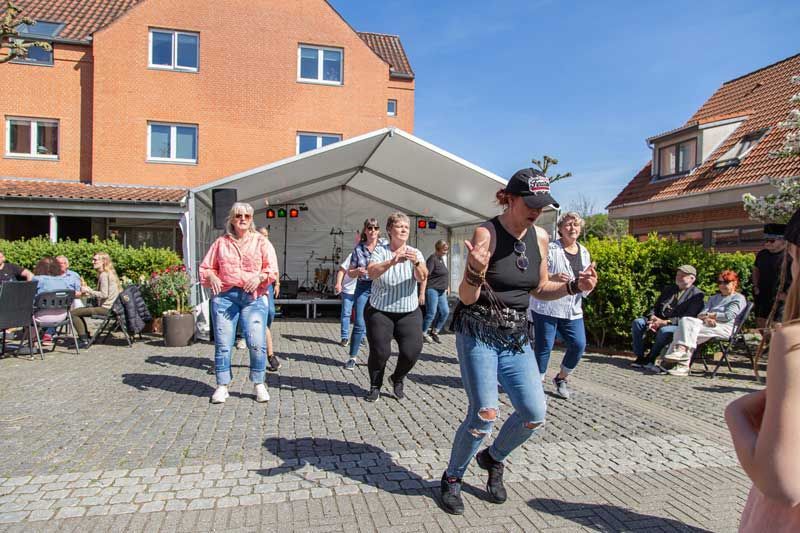 Folk danser på en murstensterrasse foran en scene opstillet under et hvidt telt; tilskuere i stole.