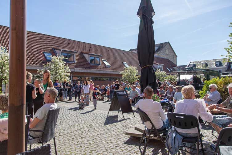 Folk samledes på en solrig udendørs plads, nogle siddende ved borde. Et band er synligt, der optræder foran publikum.