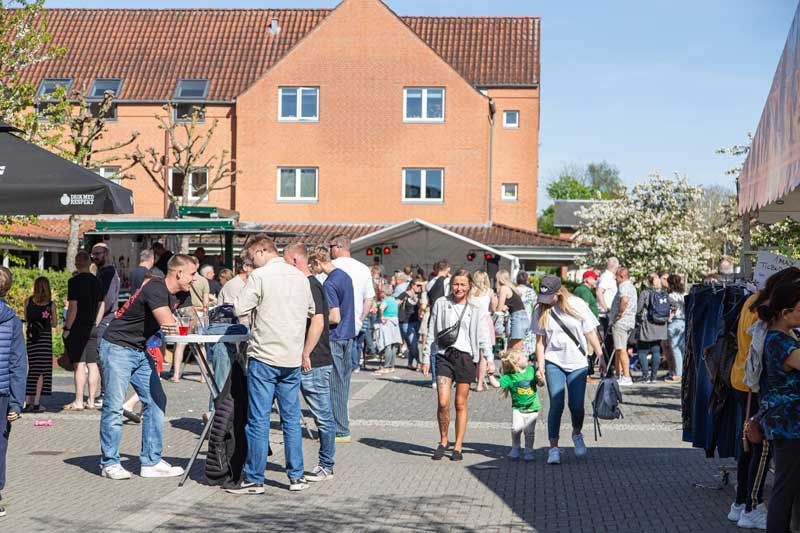 Udendørsarrangement med publikum nær en rød murstensbygning på en solskinsdag.