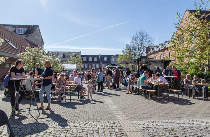 Folk ved udendørs borde, solrig gårdhave, spiser og hygger sig. Bygninger i baggrunden.