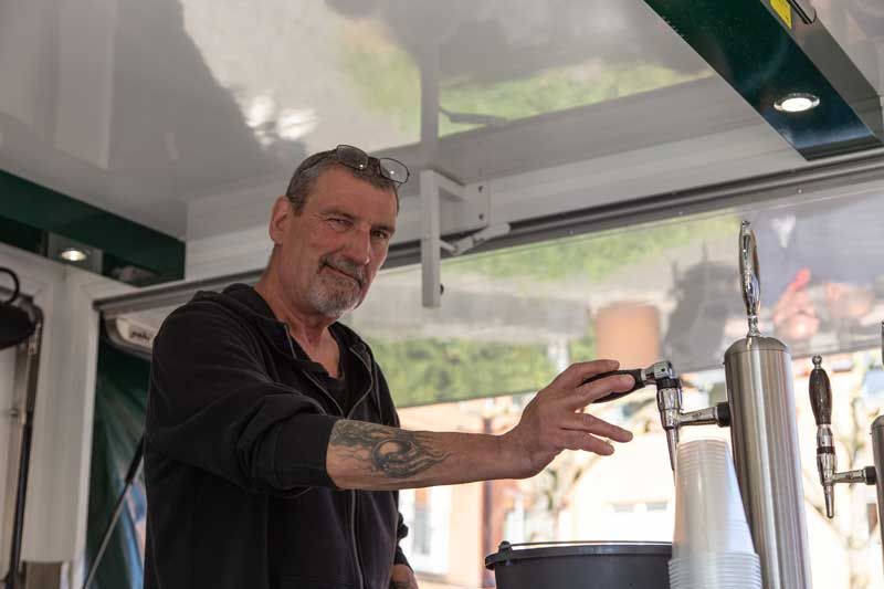 Mand hælder en drink op af hanen, smilende. Udendørs, ved en mad- eller drikkevarebod.