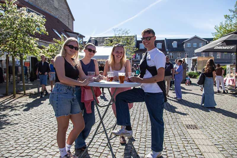 Fire personer smiler ved et bord og nyder drinks på et offentligt udendørs område på en solskinsdag.