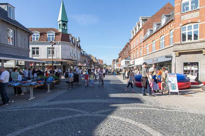 Brostensgade med mennesker, butikker og et kirketårn på en solskinsdag.
