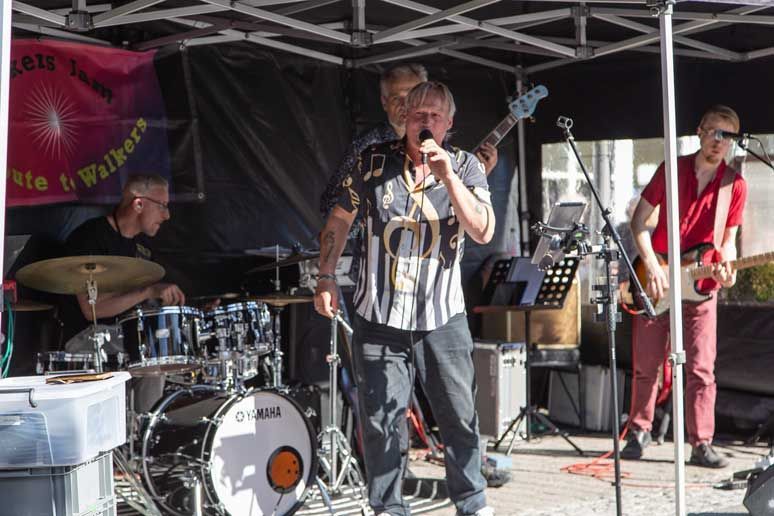 Band optræder udendørs under et baldakin: sanger, trommeslager, bassist, guitarist.