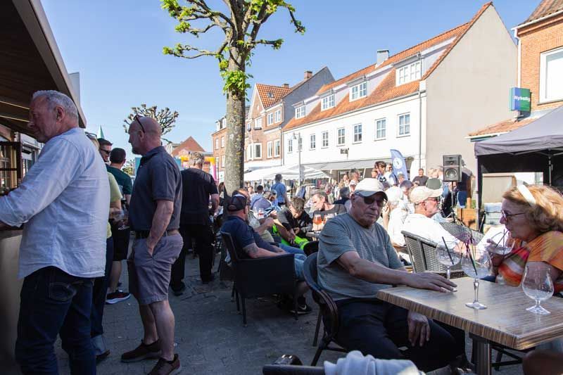 Folk på en udendørs café drikker og hygger sig. Bygninger i baggrunden under blå himmel.