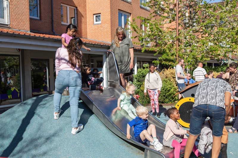 Børn leger på en metalrutsjebane på en udendørs legeplads med voksne i nærheden.