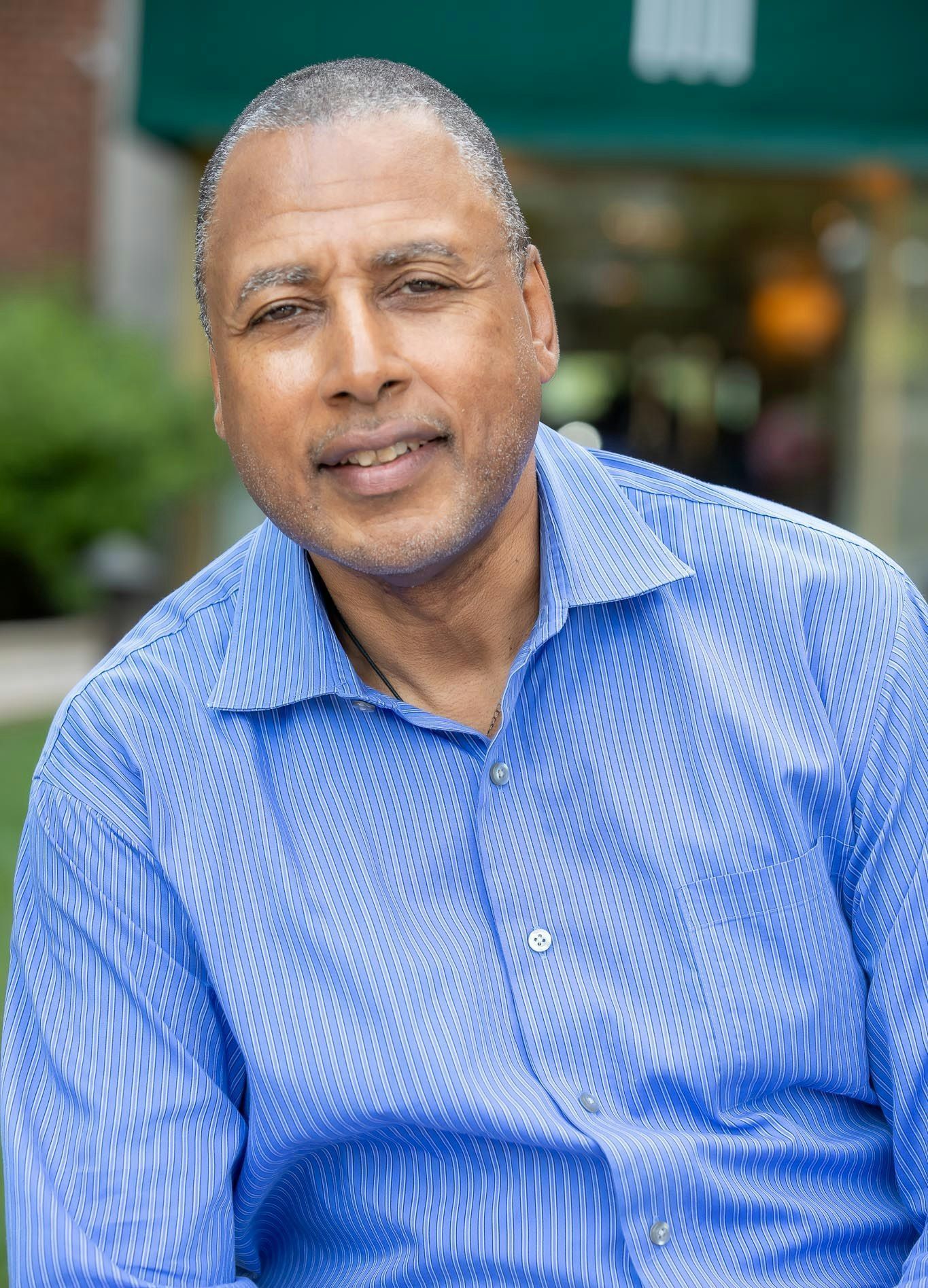 Man in blue button-down shirt smiling outdoors, blurred background of building and greenery.