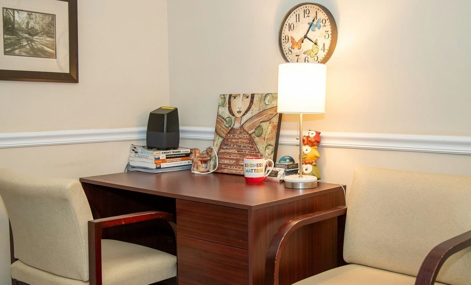 Small office corner with a desk, two chairs, a lamp, artwork, and a clock.