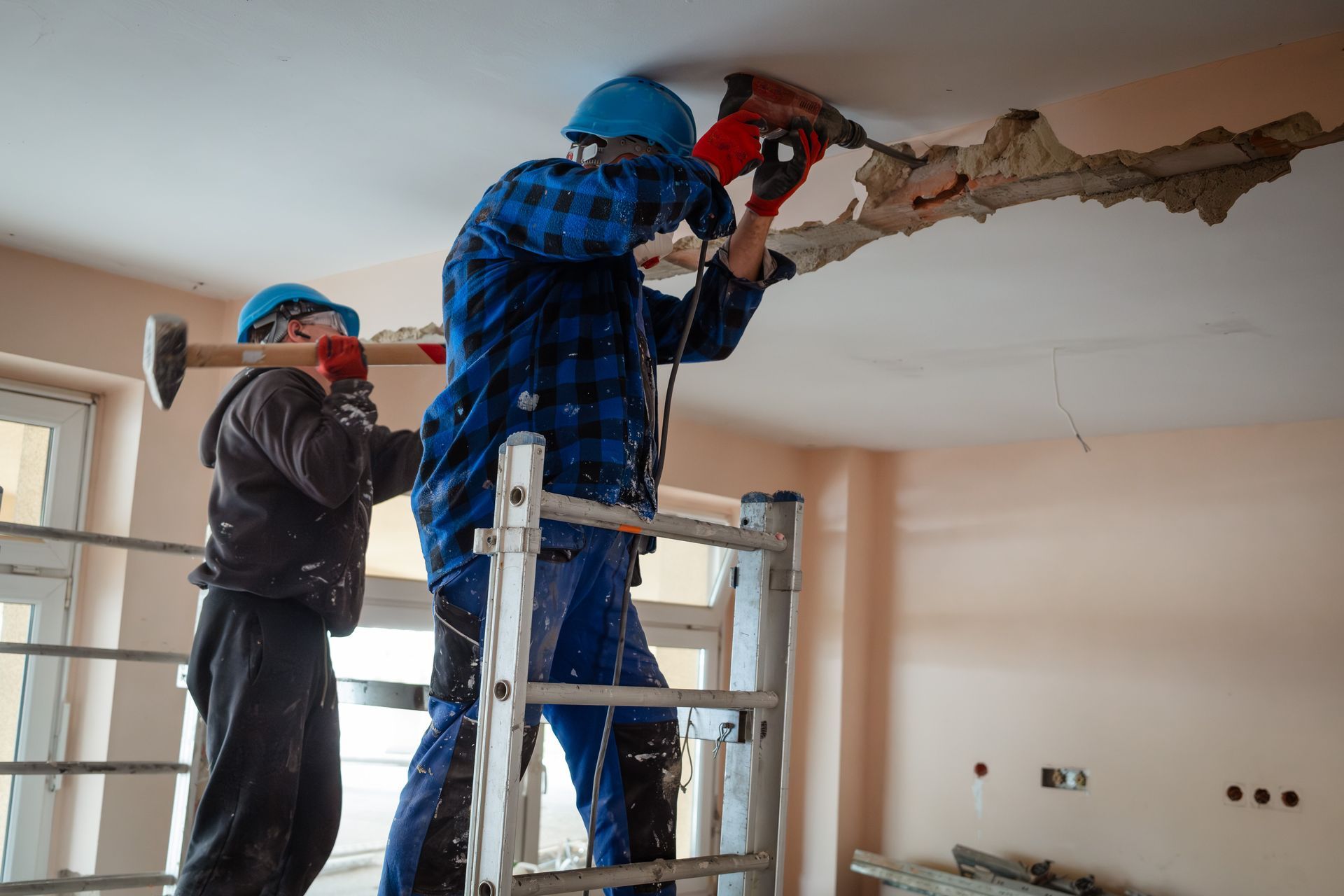 Two builders on scaffolding demolish a wall with a hammer and a drill.