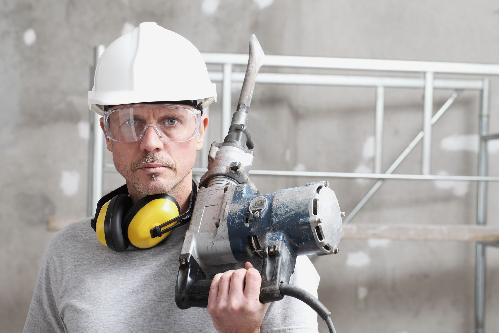 A builder with a jackhammer, wearing a hard hat, protection headphones, and protective glasses.