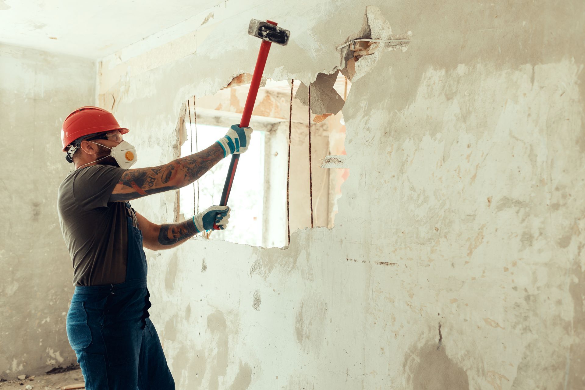 A builder dressed in a protective suit and helmet with a hammer in his hands breaks a cement wall. A builder dressed in a protective suit and helmet with a hammer in his hands breaks a cement wall.