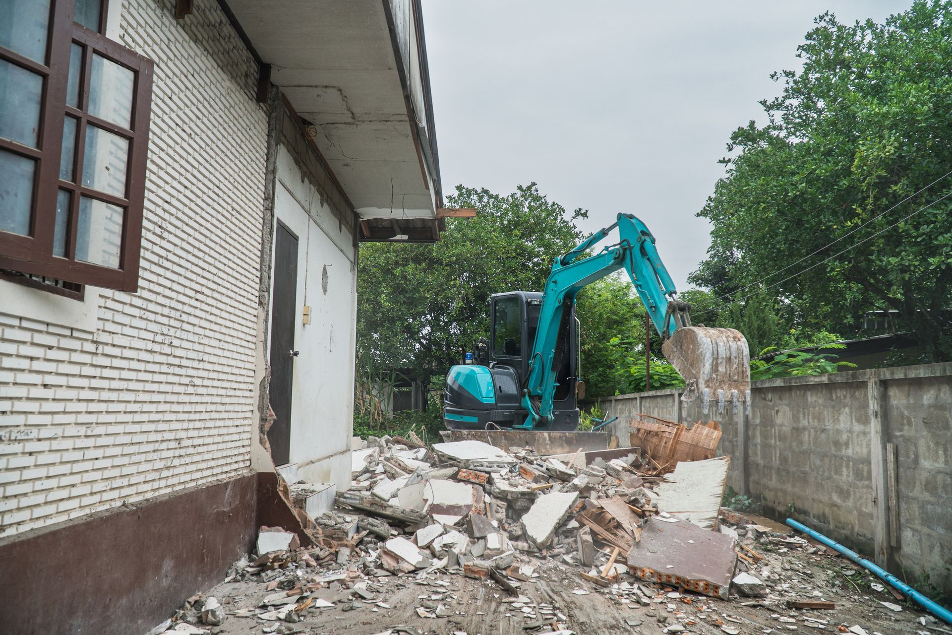 Blue excavator tearing down house wall during residential demolition with rubble on site.