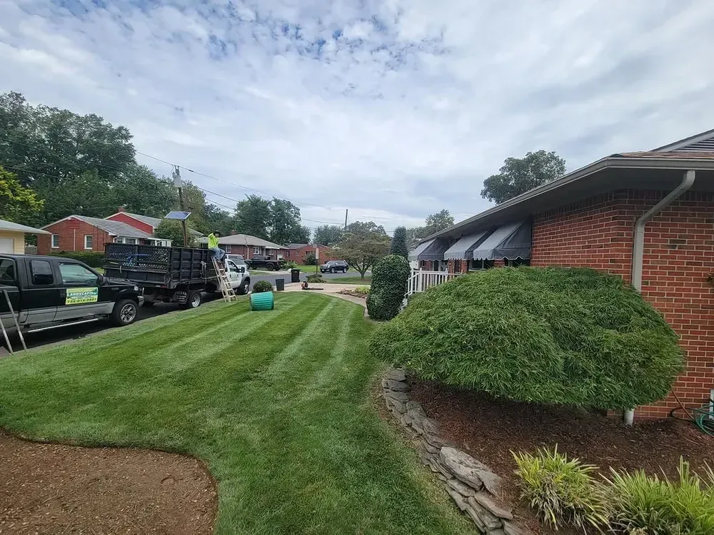 A lawn mower is cutting a lush green lawn in front of a brick house.