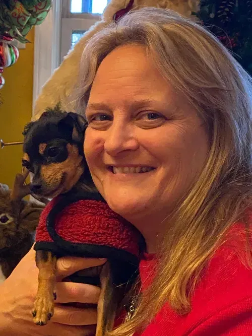 Woman with blonde hair smiles, holding a small dog in a red sweater. Festive background.
