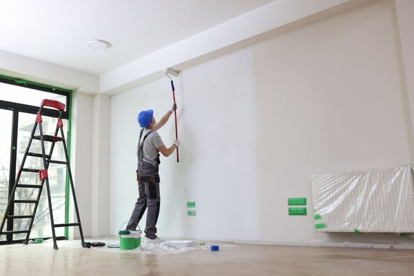 Painter in blue hard hat and overalls using a roller to paint a white wall in a room.