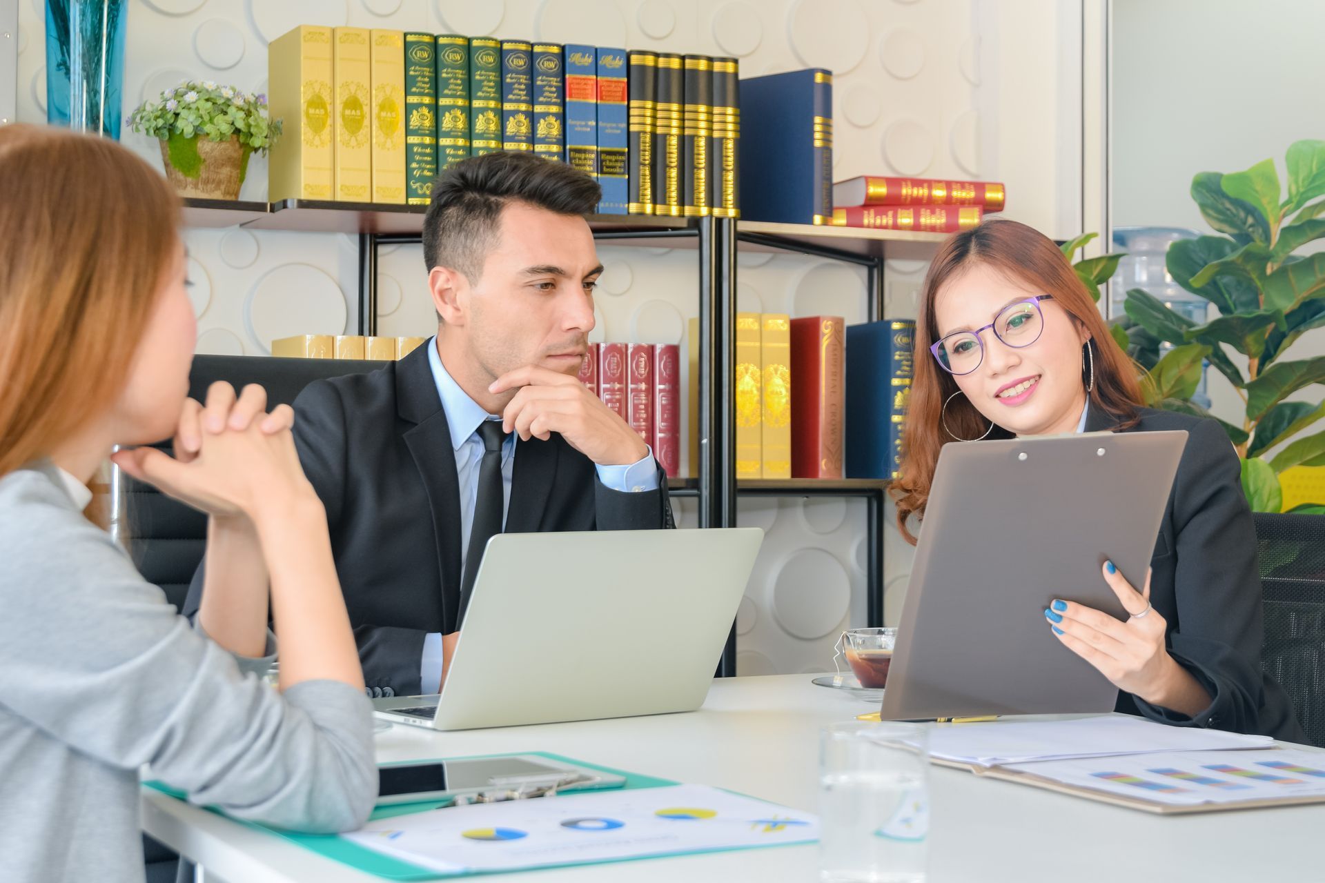 Three coworkers in a meeting around a laptop in a bright office, with one holding a tablet.