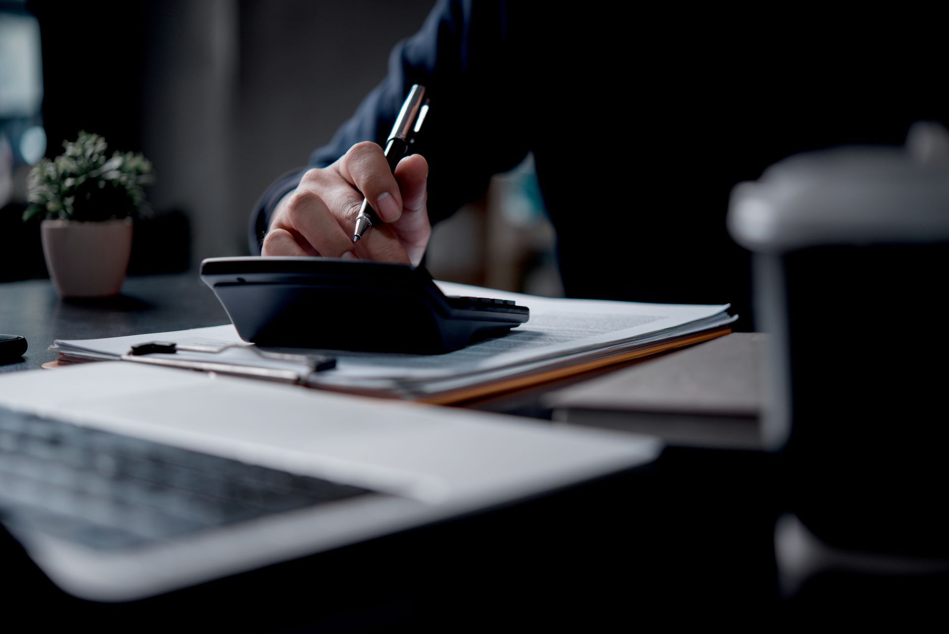 Hand writing in a notebook beside a laptop on a desk, with a small plant in the background