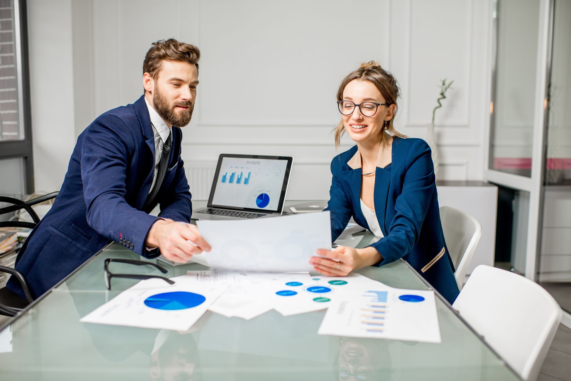 Two professionals reviewing charts at a glass conference table with a laptop in a bright office