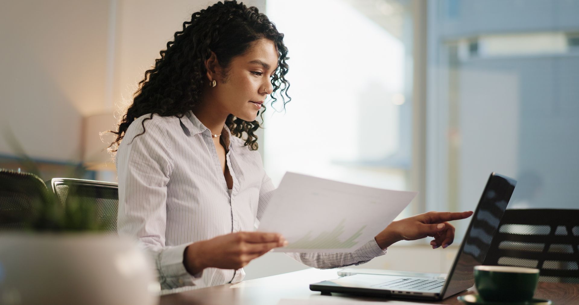 Person in a white shirt reviewing papers at a desk with a laptop in a bright office