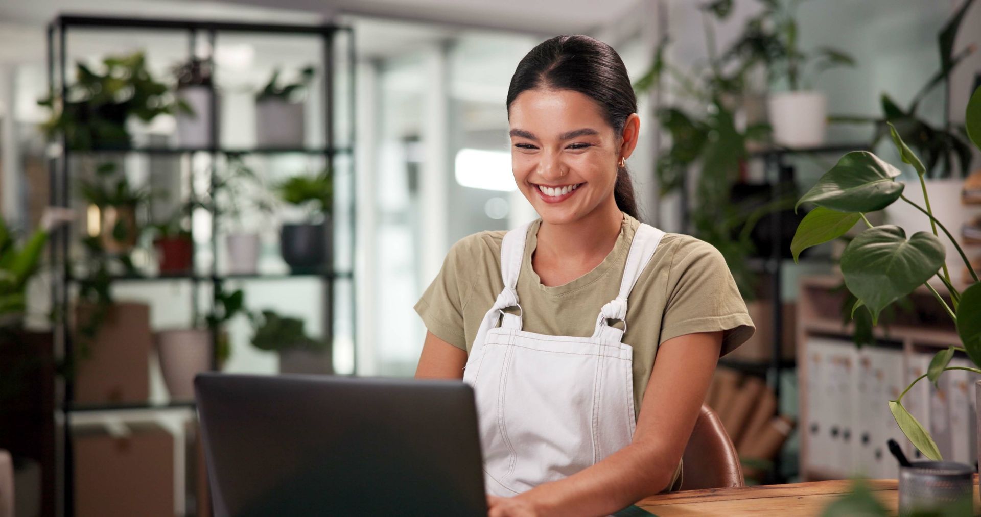 Smiling person in an apron working on a laptop in a bright plant-filled shop