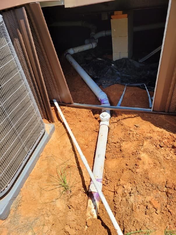 PVC pipes draining under a building foundation into the soil. Includes an AC unit and a white pipe.