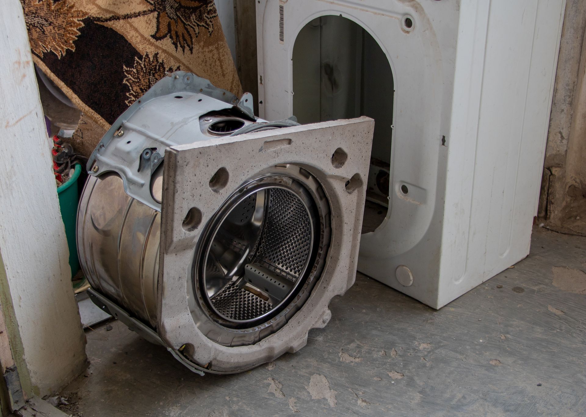 Disassembled washing machine on a concrete floor. The drum is separated from its housing.