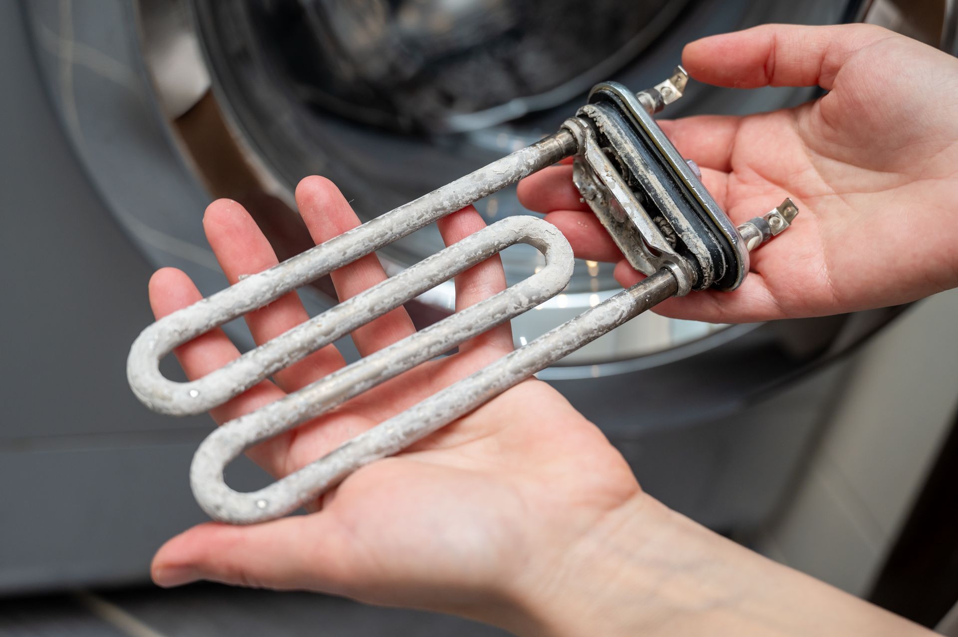 Hands holding a scale-covered washing machine heating element in front of the machine.0.0