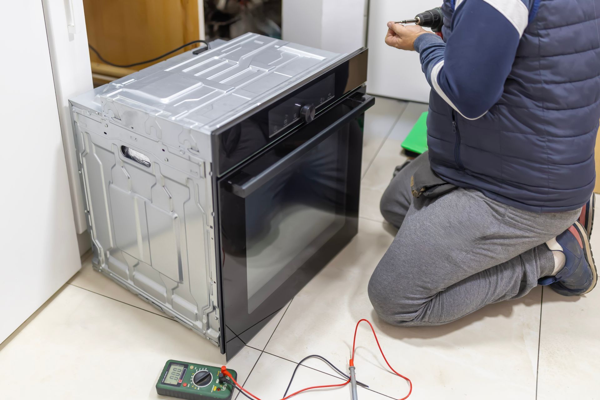 Person repairing an oven with a screwdriver; a multimeter is on the floor. Person repairing an oven with a screwdriver; a multimeter is on the floor.