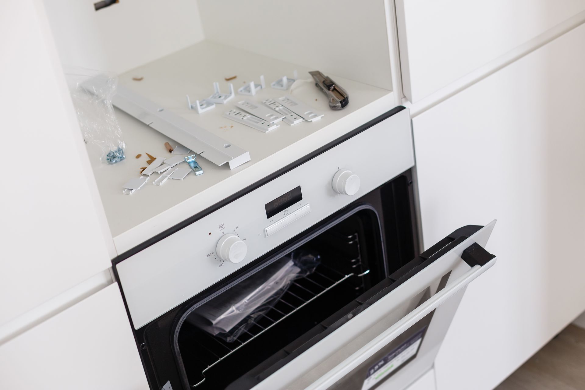 An oven installed in a white cabinet. The oven door is open. Shelf above holds hardware and a utility knife. An oven installed in a white cabinet. The oven door is open. Shelf above holds hardware and a utility knife.