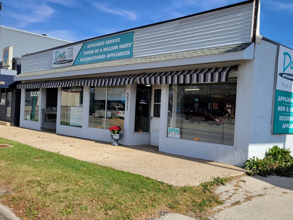 Two people standing behind a shop counter. Man in blue shirt, woman in brown, parts and supplies in background.