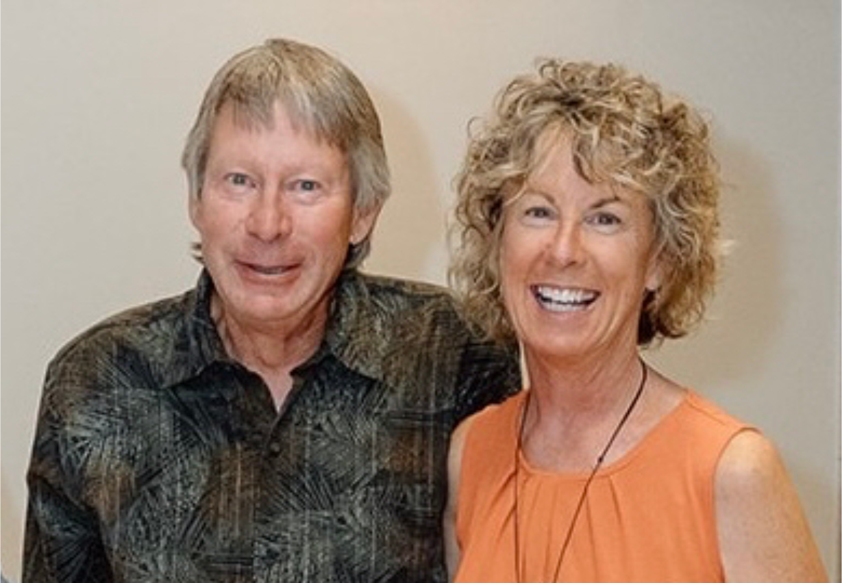 A couple poses for a photo. The woman wears an orange dress, holding a drink and purse; the man wears a patterned shirt and black pants.
