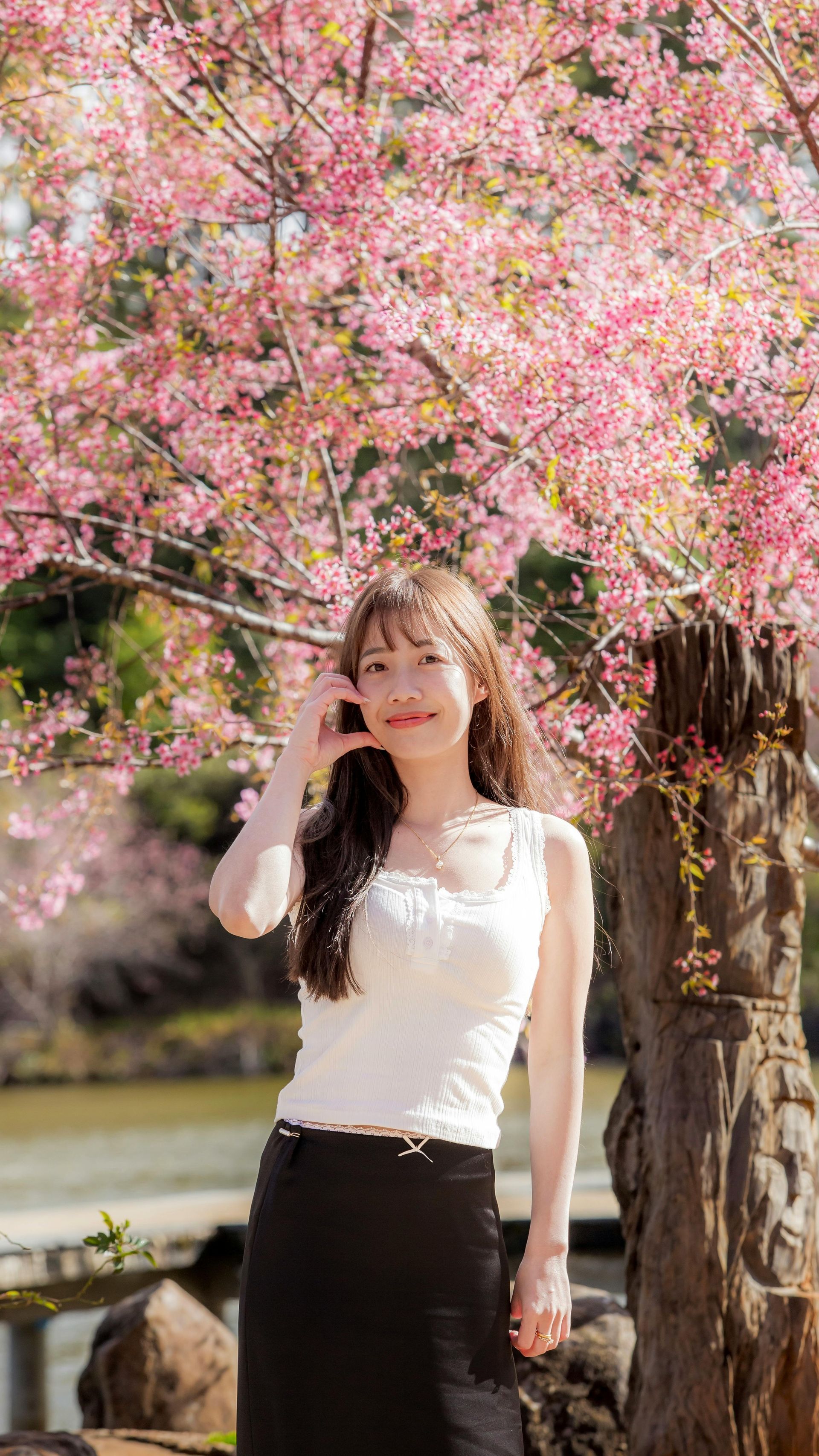 一位身穿白色上衣和黑色裙子的女子站在花園中，粉紅色的花朵簇擁著她。