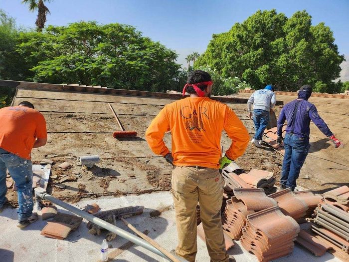 Roofers in orange and blue shirts remove tiles from a rooftop, a brush and piles of tiles nearby.