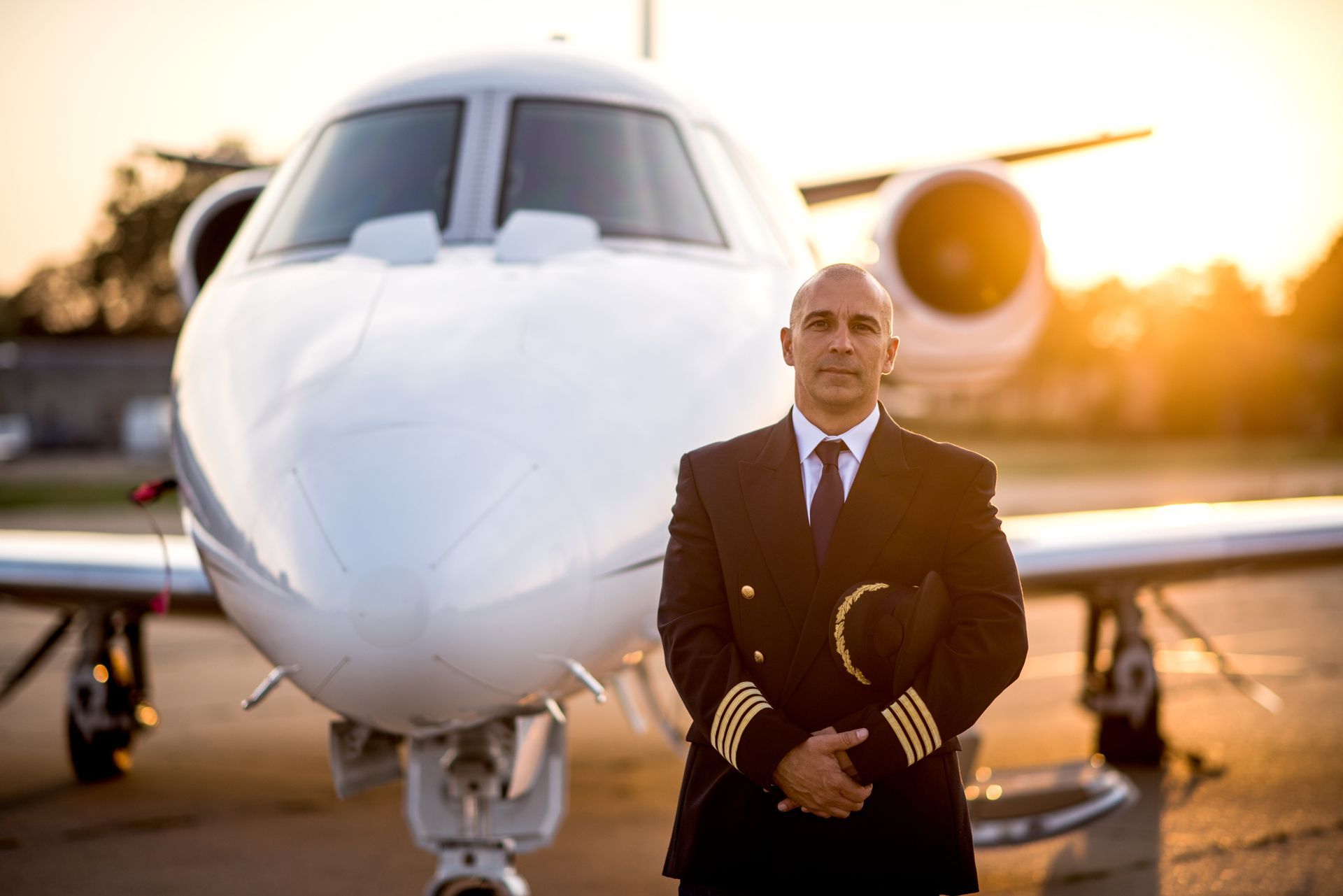 A uniformed pilot holding a cap, standing proudly by a private jet plane.