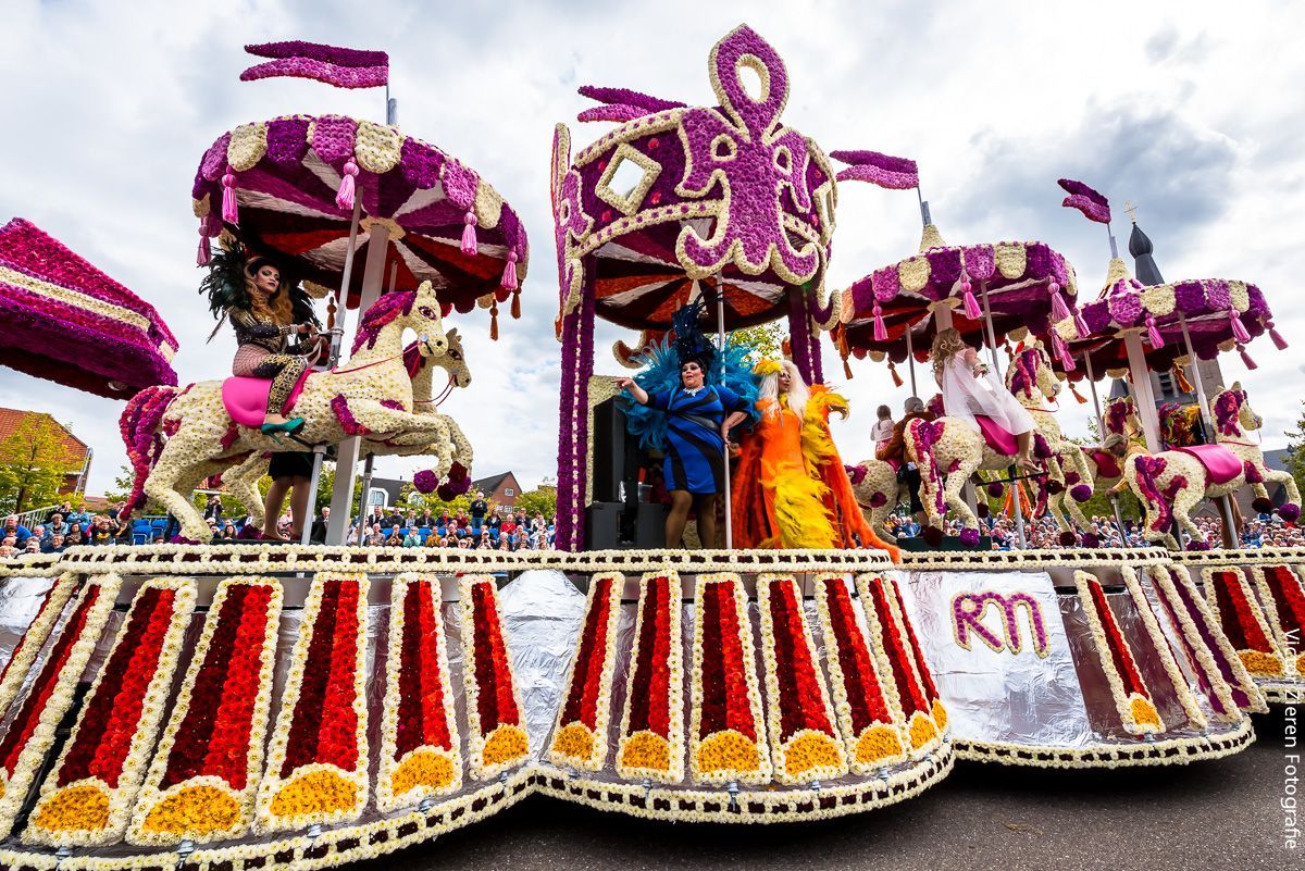 Kleurrijke festivalwagen met paarse versieringen en rood-witte draperieƫn, waarop artiesten in een parade meelopen.