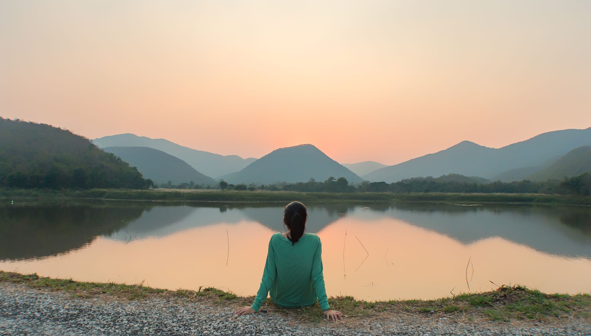 A woman is sitting on the shore of a lake at sunset.