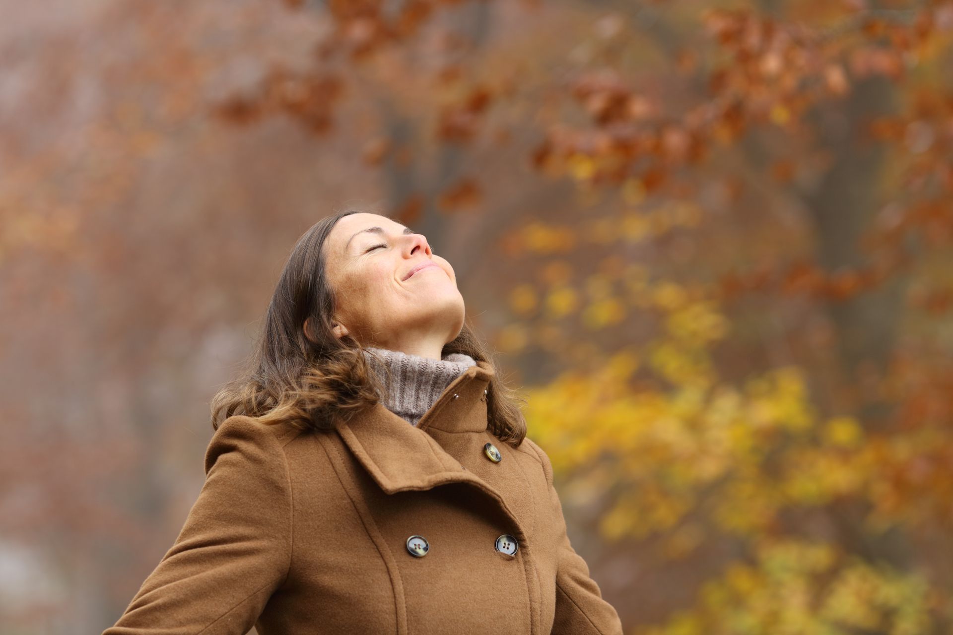 A woman in a brown coat is standing in the woods with her eyes closed.