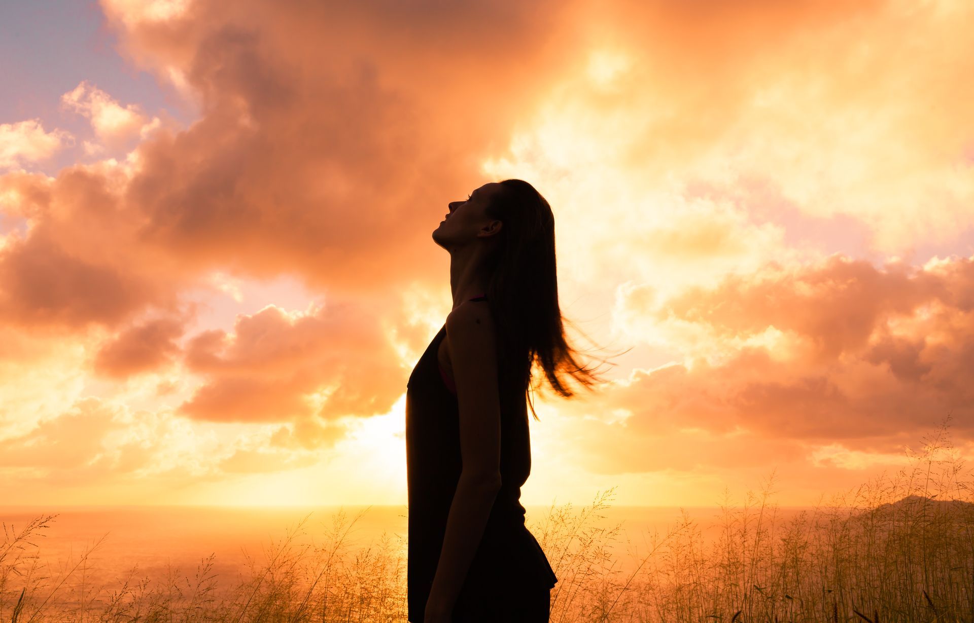 A woman is standing in a field at sunset with her eyes closed.