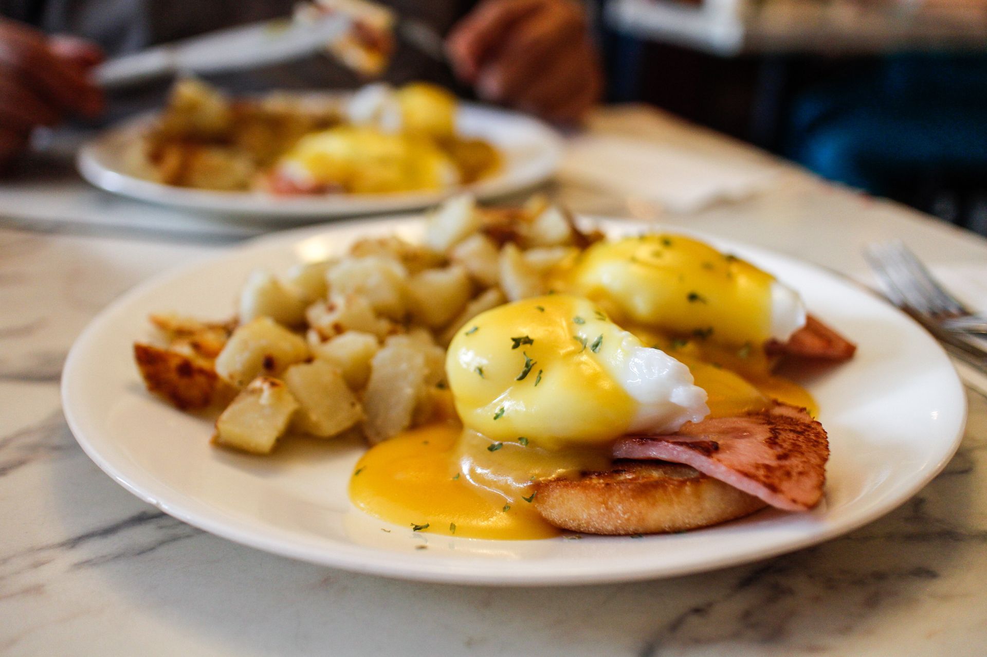 Two plates of Eggs Benedict with breakfast potatoes on a marble table.