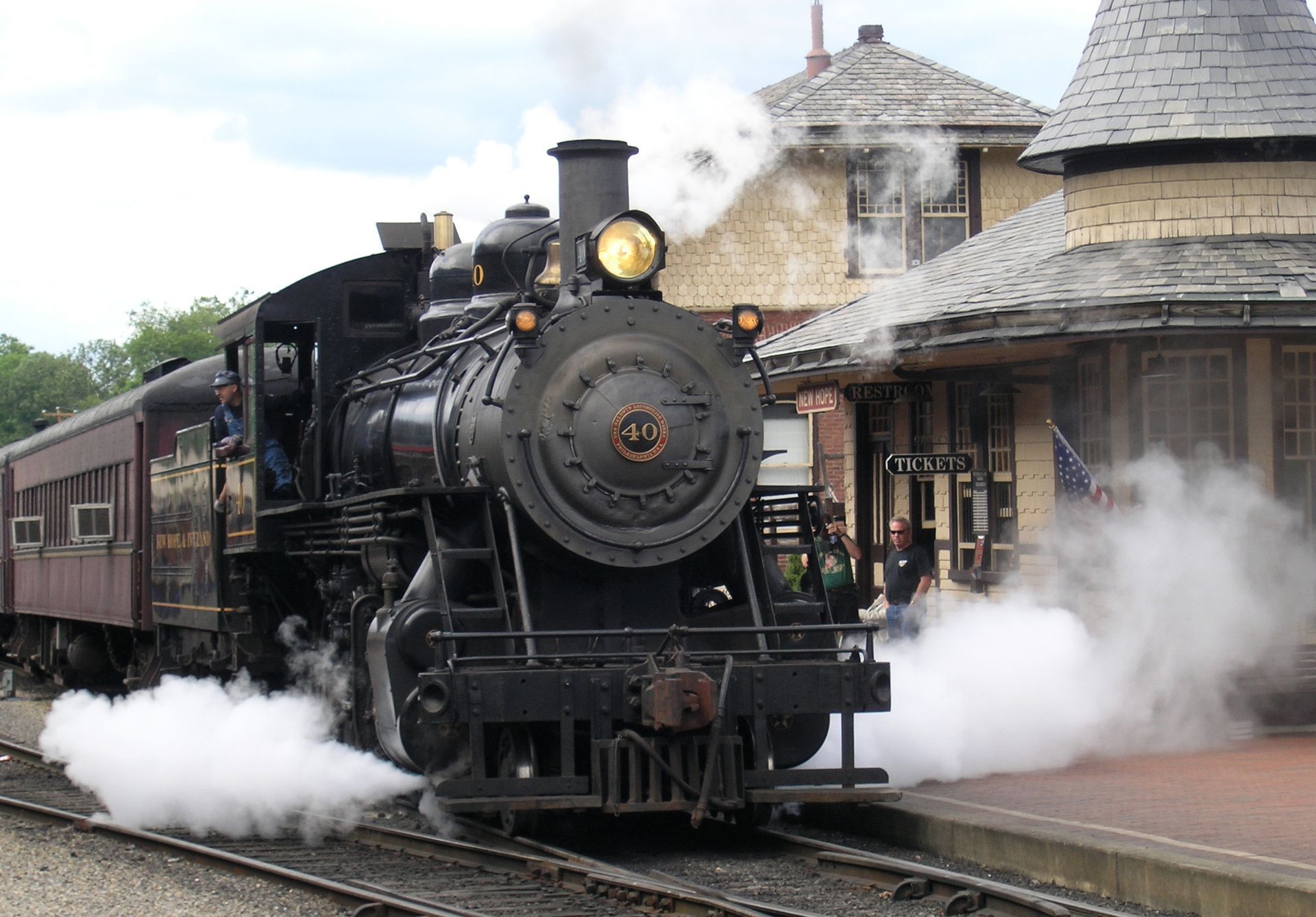 A vintage steam locomotive pulls into a historic stone station, emitting large clouds of white steam from beneath its frame.