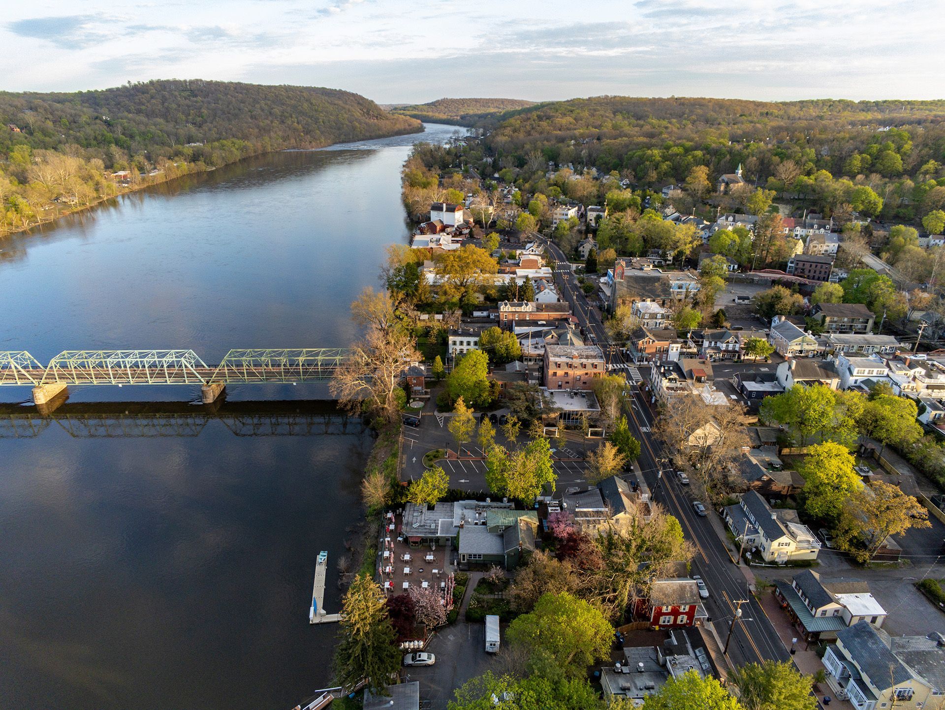An aerial view of a river flowing past a small town with a large truss bridge, surrounded by trees during autumn.