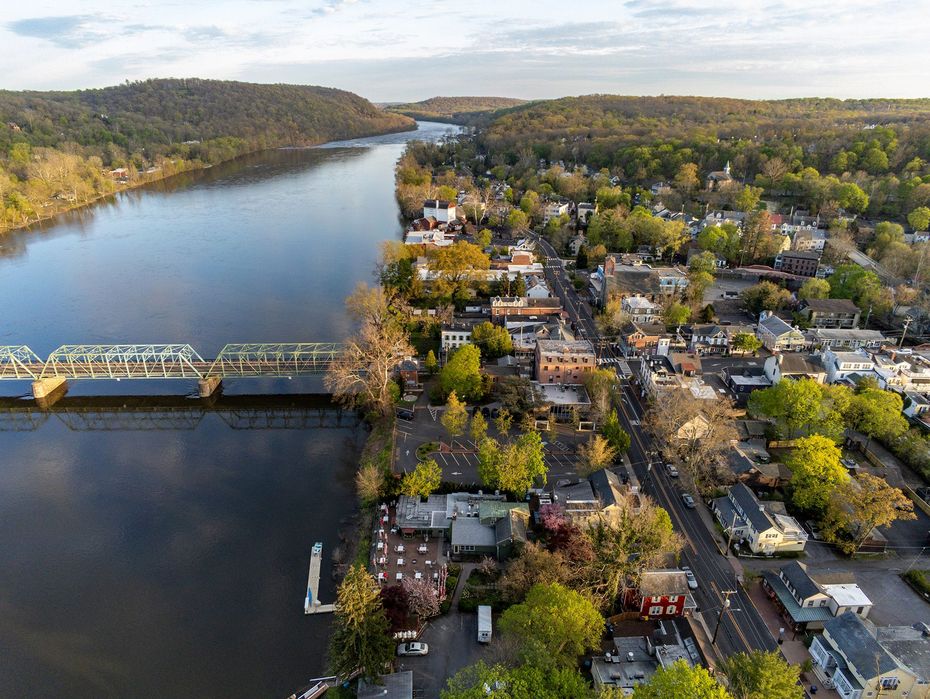 Aerial view of a small town along a river, featuring a steel truss bridge, dense trees, and buildings at sunset.